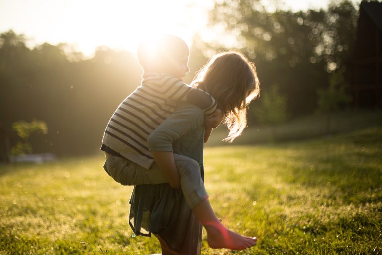 Zwei Kinder spielen draußen im Sonnenlicht. Ein Junge sitzt auf dem Rücken eines Mädchens – eine Szene, die perfekt die Bedeutung von Kinderphysio und motorischer Förderung zeigt.