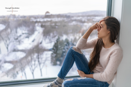 Frau sitzt nachdenklich am Fenster mit winterlicher Schneelandschaft – Symbol für Winterblues oder saisonale Beschwerden.