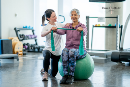 Ältere Frau trainiert mit Unterstützung einer Physiotherapeutin auf einem Gymnastikball, beide lächeln – Seniorentraining in moderner Praxisumgebung